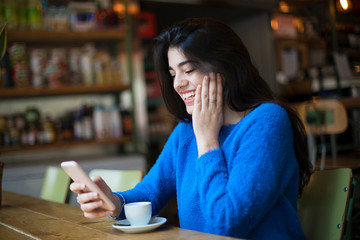 Young woman in casualwear talking through video-chat in her smartphone while sitting in cafe