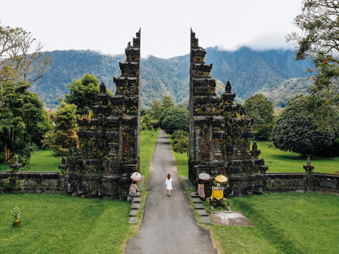 Tourist woman in white dress walking through Traditional Balinese Hindu gate Candi Bentar close to Bedugul, Bratan lake Bali island Indonesia. Vacation on Bali Drone photo