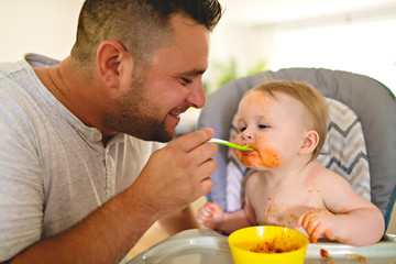 A Little baby eating her dinner and making a mess with dad on the side