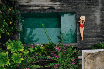 Enjoying suntan. Vacation concept. Top view of slim young unrecognizable woman in red bikini and straw hat lying near swimming pool in jungle. Back view, without face