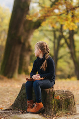 Obraz premium A little girl in black clothes is sitting on a stump and playing with autumn leaves. Blond hair girl