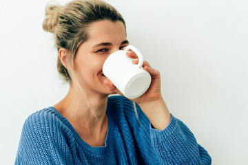 Beautiful smiling woman drinking cappuccino. Happy young woman drinking hot beverage at home. Charming blonde female holding a coffee cup. Lady wearing blue sweater sitting with cup of tea in her hand