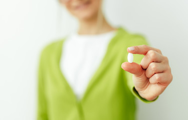 Close-up image of a pill in hand with blurred view of smiling woman. Beautiful young woman wearing green cardigan and white t-shirt taking pill isolated on white wall. Vitamin, dietary Supplements.