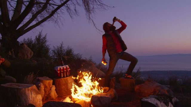 A man sprays lighter fluid on a campfire while drinking beer at a campsite.