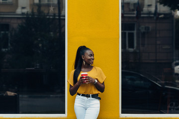Elegant black woman stands at the yellow building with coffee in her hands on the city streets