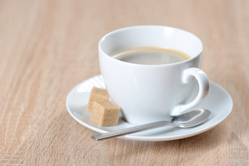 Coffee in a white cup and saucer. On a saucer are two pieces of cane sugar and a teaspoon. Light background. Free space for text.