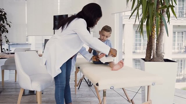 Female doctor bandaging leg of cute boy.