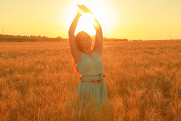 Fototapeta na wymiar girl in dress walking in golden ripe wheat field at sunset with hands upwith 