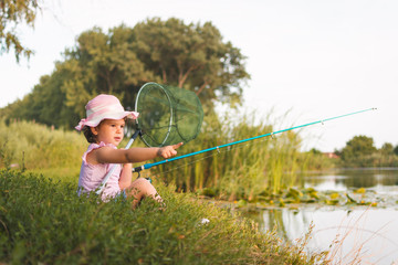 Sweet little girl sitting on the grass and patiently fishing near the river. Little girl, 4-5 years, spending fishing time alone on the river coast. Kids and fishing concept.