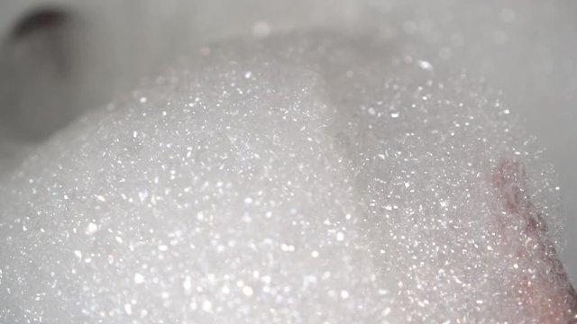 A hand raises a handful of white fresh foam in the bathroom. Bubbles sparkle in the light