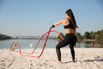 Young healthy woman doing exercise with the ropes at the beach. Single caucasian female model practicing at the river side in sunny day. Concept of healthy lifestyle, sport, fitness, bodybuilding.