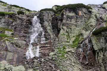 Mountain waterfall in High Tatras (Skok). Tall rocks covered with scrub. Large waterfall on the left, small water on the right. © Julian