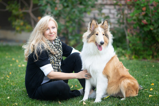 Blond Caucasian woman is sitting on a green grass and fall foliage in a park with collie dog