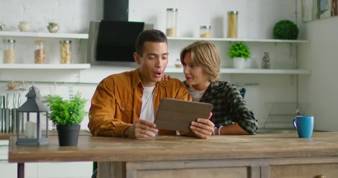 Young mixed race couple is sitting in kitchen, using tablet computer, browsing, laughing at funny photos, videos, having fun together, enjoying free time at home, Caucasian. 4K, shot on RED camera.