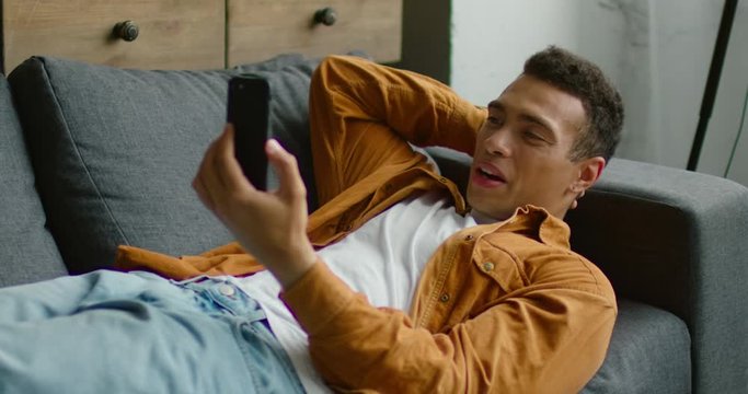 Young Hispanic man is lying on sofa, having video chat on smartphone, gadget, waving hello, smiling, in good mood, enjoying conversation with his friends, family. Slow motion, 4K. Shot on RED camera.