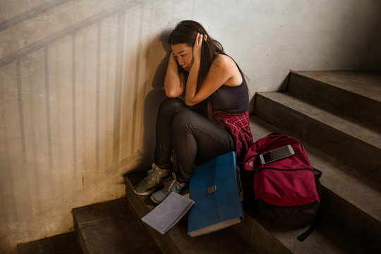 Dramatic portrait of Asian female college student bullied. Young depressed and sad Japanese girl sitting lonely on campus staircase suffering bullying and harassment