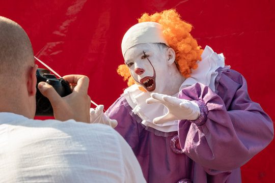 A man with a camera photographs the face of a very scary clown with red hair and sharp teeth close-up. A teenager in a suit IT opened his mouth. Cosplay for carnival or for halloween.
