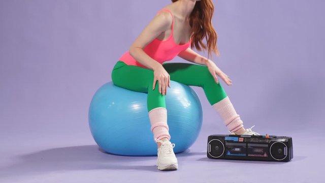 Redhead girl pressing the button on the portable radio cassette player to turn on music to start an active aerobics workout. sitting on a blue fit ball in leggings and training shoes