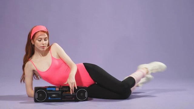 Fit ginger female model poses for commercial photoshoot for aerobics sportswear and shoes, sits on floor with hand on portable audio player, perfect curves and sporty body shape, wears pink bodysuit