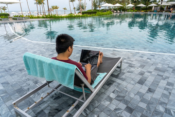 Asian man using laptop on chair at beautiful swimming pool 