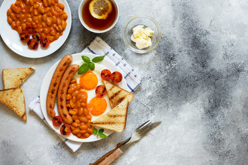 English breakfast with fried sausages, beans, mushrooms, fried eggs, grilled cherry tomatoes. Served with a cup of tea with lemon, bread toast and butter. Gray background, top view, copyspace