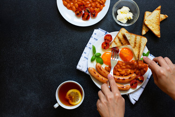 Female hands as she is having English breakfast with fried sausages, beans, mushrooms, fried eggs, grilled tomatoes. Served with a cup of tea with lemon, bread toast and butter. Black background