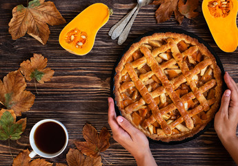 Female hands holding homemade pumpkin pie. Halloween and Thanksgiving. Holiday pumpkin sweets. Wooden autumn background, dry leaves, cut pumpkin, cup of tea. Top view. Copyspace. 
