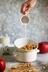 Granola with nuts and raisins in a white bowl. Healthy breakfast served on a light table with red apples. Milk is pouring into a bowl from a cup. Female hand is pouring milk. Vertical