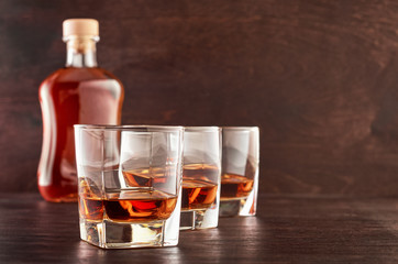 Three glasses of whiskey on a wooden table, a full bottle of whiskey in the background