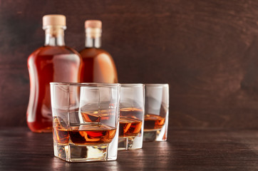 Three glasses of whiskey on a wooden table, two full bottles of whiskey in the background