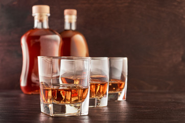 Three glasses of whiskey on ice on a wooden table, two full bottles of whiskey in the background
