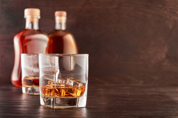Two glasses of whiskey with ice on a wooden table, in the background two bottles of whiskey of...