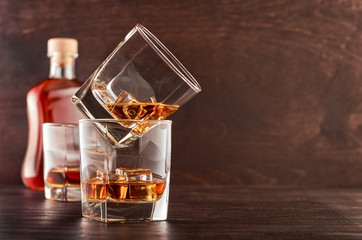 Two glasses of whisky with ice on a wooden table. One glass stands on the other. In the background, a full bottle of whiskey and a glass of whiskey