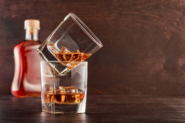 Two glasses of whisky with ice on a wooden table. One glass stands on the other. In the background is a full bottle of whiskey
