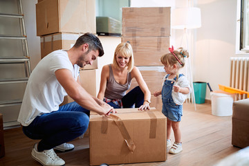 Smiling parents with child moving in new home unpacking boxes.