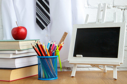 School supplies, miniature chalk board, a stack of books and an apple on the table against the background of a white shirt with a tie on a hanger, soft focus