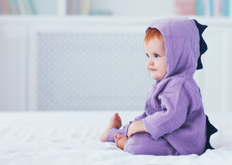 beautiful smiling, nine month old, infant baby girl in dinosaur costume, is sitting on the bed