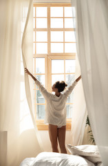 Fototapeta na wymiar Young woman in shirt opening curtains in the morning