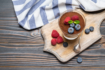 Classic tiramisu dessert portion with blueberries and strawberries in a glass cup on cutting board on wooden background