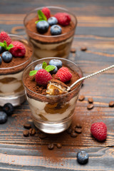 spoon with piece of Classic tiramisu dessert with blueberries and strawberries in a glass cup on wooden background