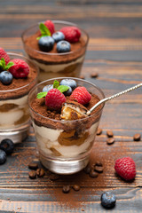 spoon with piece of Classic tiramisu dessert with blueberries and strawberries in a glass cup on wooden background