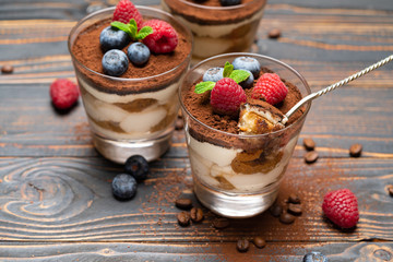 spoon with piece of Classic tiramisu dessert with blueberries and strawberries in a glass cup on wooden background