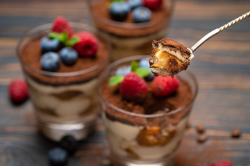 spoon with piece of Classic tiramisu dessert with blueberries and strawberries in a glass cup on wooden background