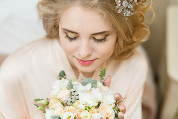 Pretty young girl. Blonde woman with luxurious long curly hair. Boudoir morning of the bride. Taking wedding bouquet in her hands. Touching her neck