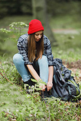 Female hiker wearing red hat tying shoelaces outdoors in the forest, near backpack
