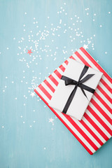 Fototapeta premium Top view of gifts lying on a blue wooden background. Stylish striped gift and a gift wrapped in white paper tied with a black ribbon. Silver stars are scattered on the table like confetti.