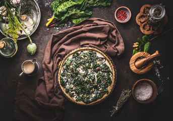 Quiche or open faced savory pie cooking preparation on dark rustic background with kitchen utensils and ingredients, top view. Tasty seasonal baking