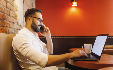 Handsome bearded man sitting in a coffee shop and working for a laptop while talking on the smartphone
