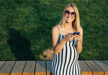 Photo of pregnant blonde woman in long dress with phone in her hand standing near lake