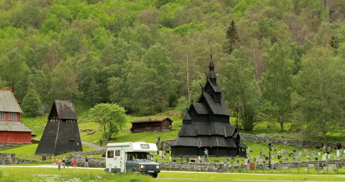 Borgund, Norway. Caravan Motorhome Car Goes On Road Near Famous Norwegian Landmark Stavkirke An Old Wooden Triple Nave Stave Church In Summer Day. Panorama, Panoramic View